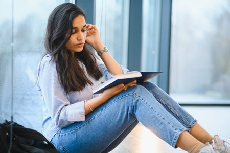 Indian student sitting by window reading a book, focusing on education and acquiring knowledge in a study environmentの写真素材