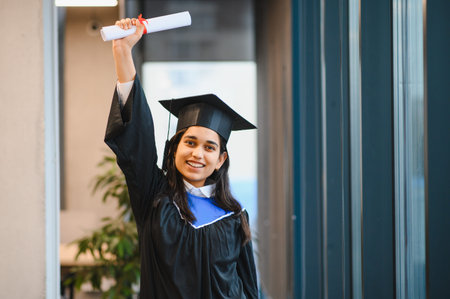 Young Indian woman wearing academic gown and cap, smiling and holding her diploma, celebrating academic achievementの写真素材