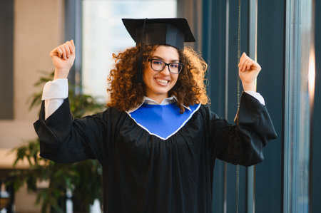Female student wearing graduation cap and gown, smiling confidently and raising arms in celebration of academic achievementの写真素材