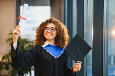 Young female graduate student smiling proudly while holding her diploma and graduation cap after completing university studiesの写真素材