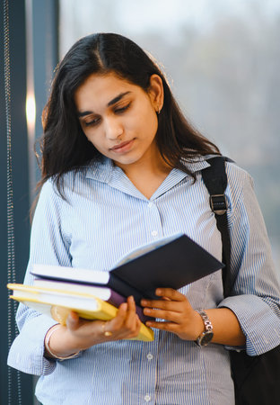 Young Indian woman wearing a backpack and striped shirt, deeply engrossed in reading books while standing indoorsの写真素材