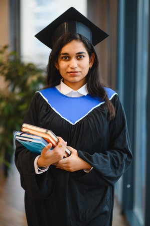 Young Indian woman celebrating academic success, wearing a mortarboard and gown, holding study books in a university settingの写真素材