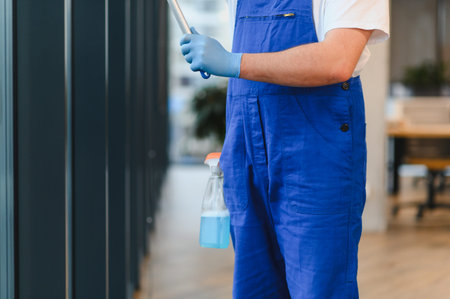 Professional cleaner at work, wearing protective gloves and overalls, cleaning windows with a squeegee and spray bottleの写真素材