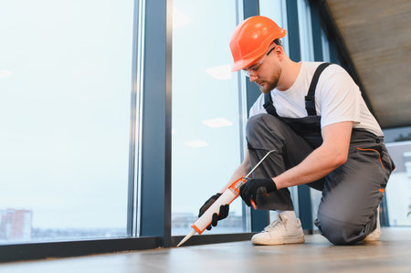 Worker in safety gear sealing window gap with a caulking gun. Highlighting maintenance, repair, and...