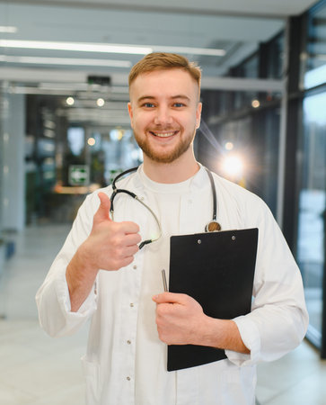 Young male doctor in white coat with stethoscope and clipboard, smiling and giving thumbs up in...