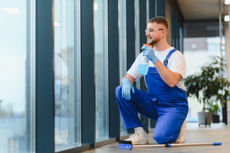 Cleaning worker in blue uniform and safety glasses spraying cleaner on large windows, providing...