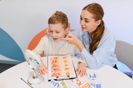 Speech therapist assisting a young boy with speech development using a mirror and articulation cards at a tableの写真素材