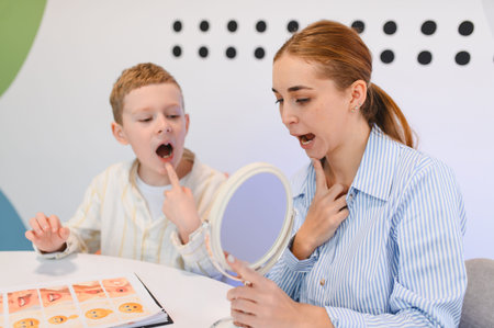 Boy performing articulation exercises with a speech therapist using a mirror to observe mouth...