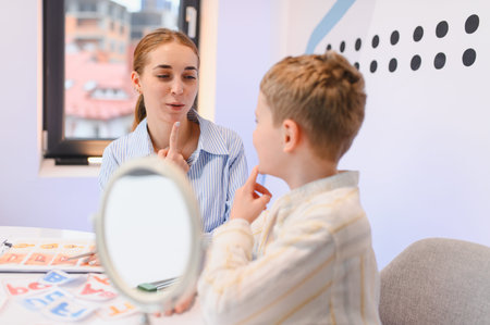 Qualified speech therapist working with a young boy, pointing to her mouth while he imitates,...