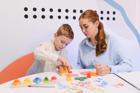 Young boy playing with wooden educational toys while a female teacher assists him in a bright...