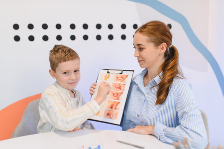 Boy pointing at a visual chart representing various mouth expressions during a speech therapy session with his female therapistの写真素材