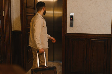 Man holding luggage and waiting for an elevator to arrive in a hotel lobby, preparing for travel or departureの写真素材