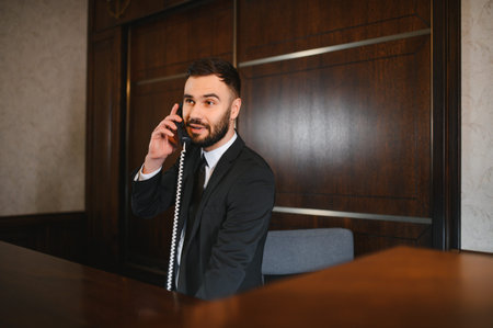 Male receptionist manning hotel front desk, talking on telephone, providing customer service in a professional lobbyの写真素材