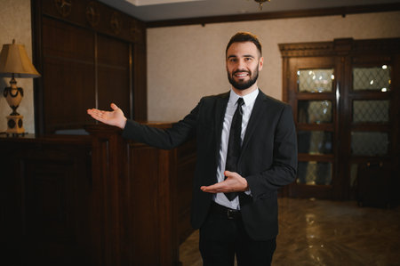 Smiling bearded man working as a receptionist or concierge, welcoming guests in a luxurious hotel lobby, offering hospitality servicesの写真素材