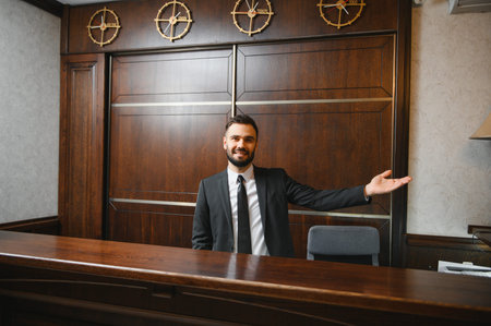 Friendly receptionist in a suit standing at a wooden desk, offering service and hospitality in a hotel lobbyの写真素材