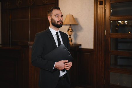 Man in a black suit holding a clipboard, standing in a classic hotel lobby. Professional male at reception areaの写真素材