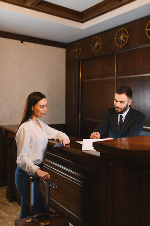 Woman guest with luggage checking in at hotel reception desk. Male receptionist assisting a traveler. Hospitality serviceの写真素材