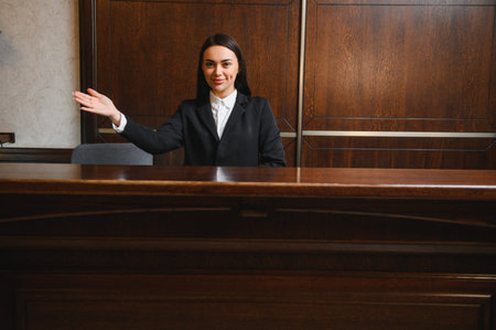 Friendly woman receptionist in uniform smiling and gesturing, providing service at a hotel or office reception areaの写真素材