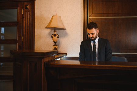 Male receptionist wearing a suit and tie working at a wooden reception desk in a hotel lobby, looking downの写真素材