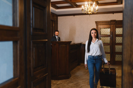 Woman guest carrying suitcase walking into a hotel lobby, smiling at the camera. Male receptionist working behind the counterの写真素材
