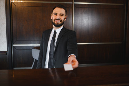 Young hotel receptionist in suit smiling at guest while handing over a key card at the modern lobby front desk during check inの写真素材