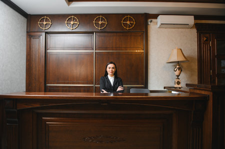 Friendly female receptionist smiling at the camera, providing service at a luxurious hotel front desk, ready to assist guestsの写真素材