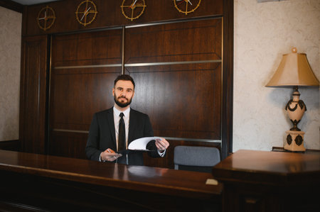 Young man concierge standing behind the reception counter, holding documents and pen for guest check inの写真素材