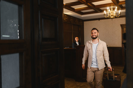 Man with suitcase entering a hotel lobby, being greeted by a receptionist. Concept of travel, hospitality, and customer serviceの写真素材