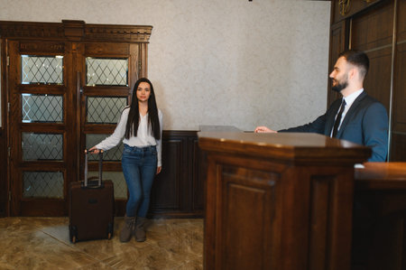 Woman guest carrying luggage arriving at a hotel, standing near the reception desk with hotel employee assistingの写真素材