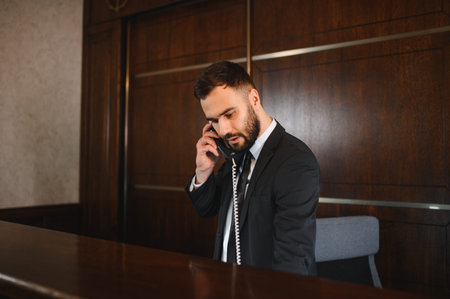 Hotel receptionist man using landline phone at the reception desk in a lobby, providing service to guestsの写真素材