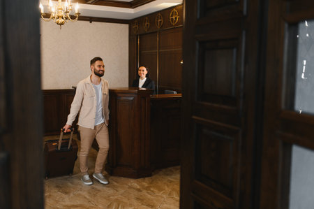 Male traveler entering a luxurious hotel lobby, smiling at the front desk receptionist while holding his luggageの写真素材
