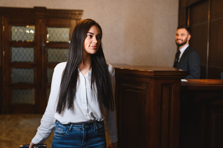 Young woman standing in a hotel lobby with a smiling male receptionist in the background, ready for travel and accommodationの写真素材