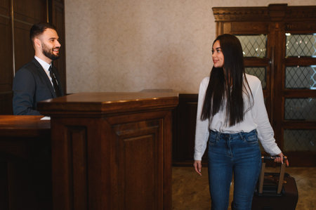 Male receptionist smiling at a female guest arriving with luggage, checking in at a hotel reception deskの写真素材