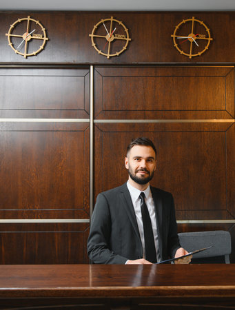 Friendly male receptionist in black suit working at luxury hotel lobby desk, with various time zone clocks hanging aboveの写真素材
