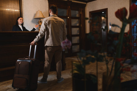 Man with suitcase entering hotel lobby, smiling receptionist welcoming guest at front desk for check in processの写真素材