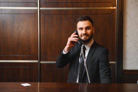 Male receptionist smiling, talking on a landline phone. Standing in a hotel lobby, wearing a professional suit, providing serviceの写真素材