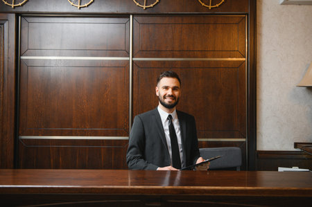 Friendly receptionist in suit holding a clipboard, standing at the reception desk, ready to assist guestsの写真素材