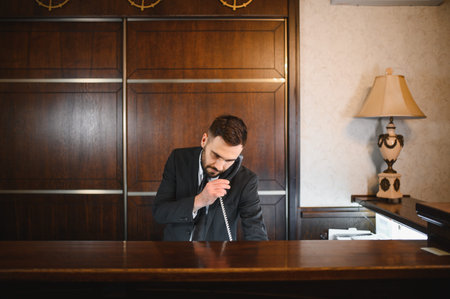 Male receptionist helping a guest by phone at a hotel lobby desk, providing excellent customer service and welcoming atmosphereの写真素材