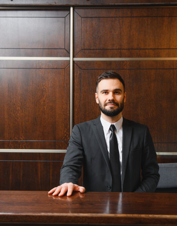 Friendly male receptionist standing behind counter, providing professional hospitality service in a hotel lobby interiorの写真素材
