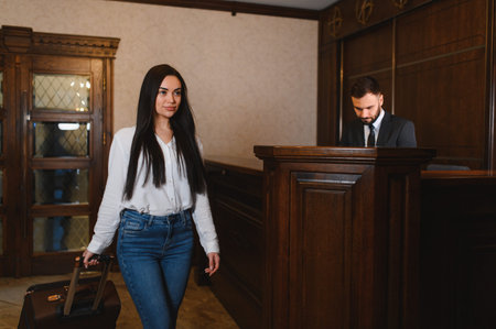 Woman traveler carrying luggage arriving at a hotel lobby, approaching the reception desk with a male receptionistの写真素材