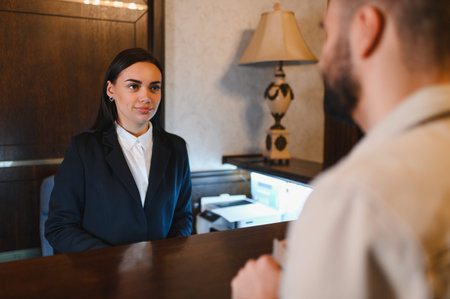 Friendly female hotel receptionist standing behind a counter, providing service to a male guest...