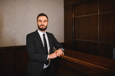 Professional man wearing a black suit and tie, posing at a wooden reception counter in a hotel lobbyの写真素材