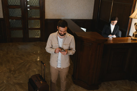 Traveler standing with luggage in a hotel lobby, smiling and using a mobile phone while a female receptionist works at the counterの写真素材