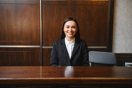 Young professional woman providing excellent hospitality service at a hotel reception desk, looking at camera and smilingの写真素材