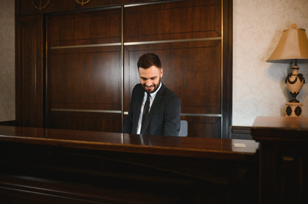 Friendly male receptionist in a suit looking down and smiling behind a wooden reception desk in a hotel lobbyの写真素材