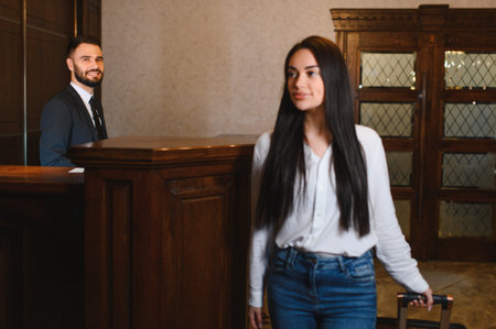 Female guest arriving at a hotel, walking through the lobby with her suitcase while male concierge is smiling behind the receptionの写真素材