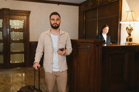 Young man with luggage and smartphone getting ready for check in at a classic hotel lobby with receptionist in backgroundの写真素材
