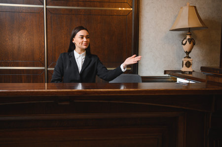 Young professional woman standing at luxurious hotel reception desk, welcoming and offering assistance with a smileの写真素材