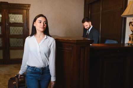 Young woman guest with luggage checking into hotel lobby. Male receptionist working at front desk in backgroundの写真素材
