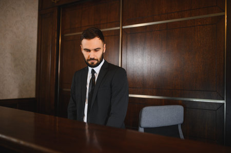 Professional man in a suit standing behind a reception desk in a hotel lobby, providing attentive serviceの写真素材